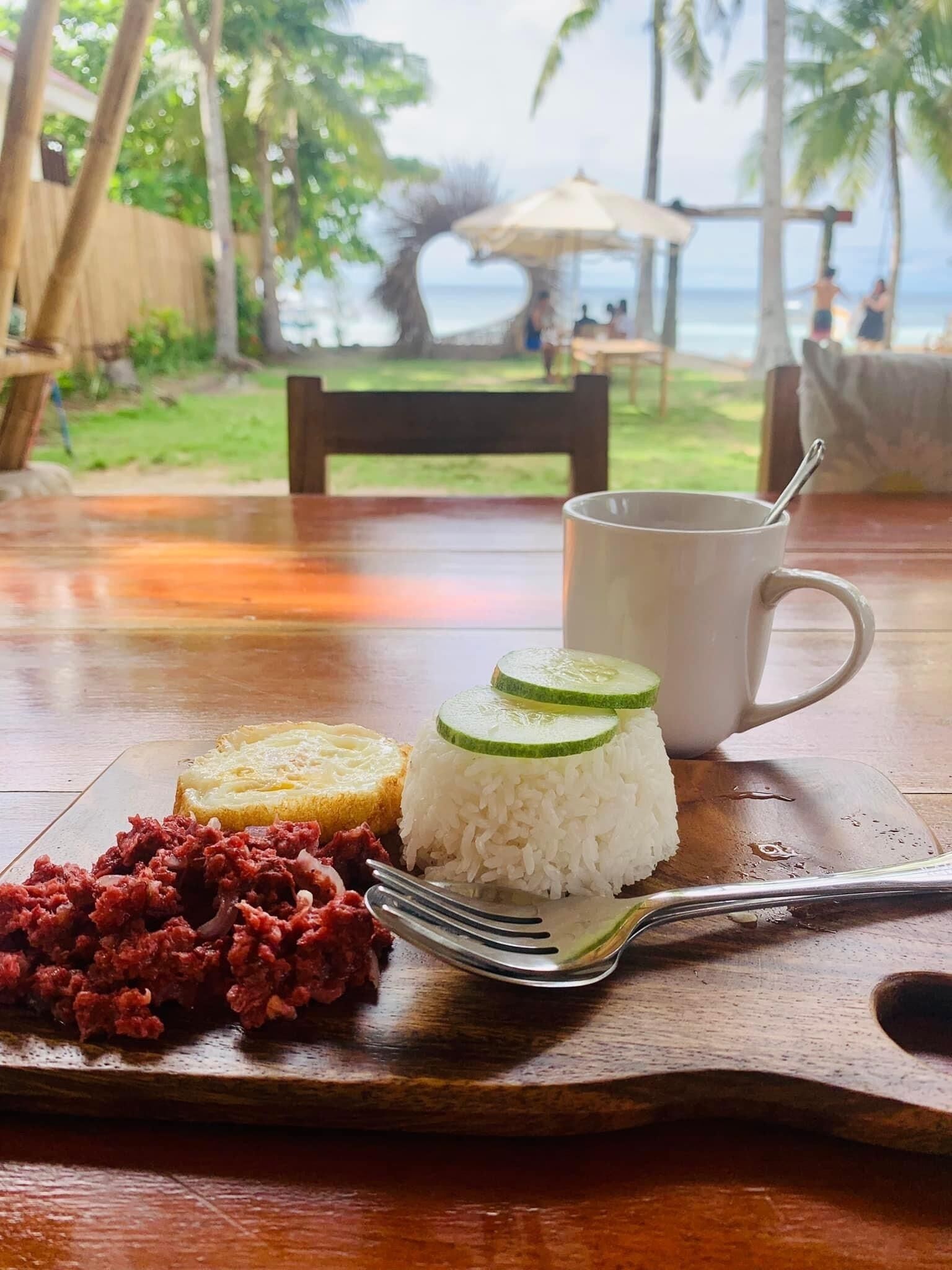 A wooden table with a meal consisting of rice, cucumber slices, and a meat dish, accompanied by a white mug and a fork.