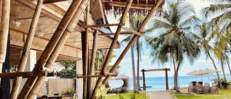 Un espacio exterior techado con estructuras de bambú, plantas en macetas y vistas a la playa y al océano.