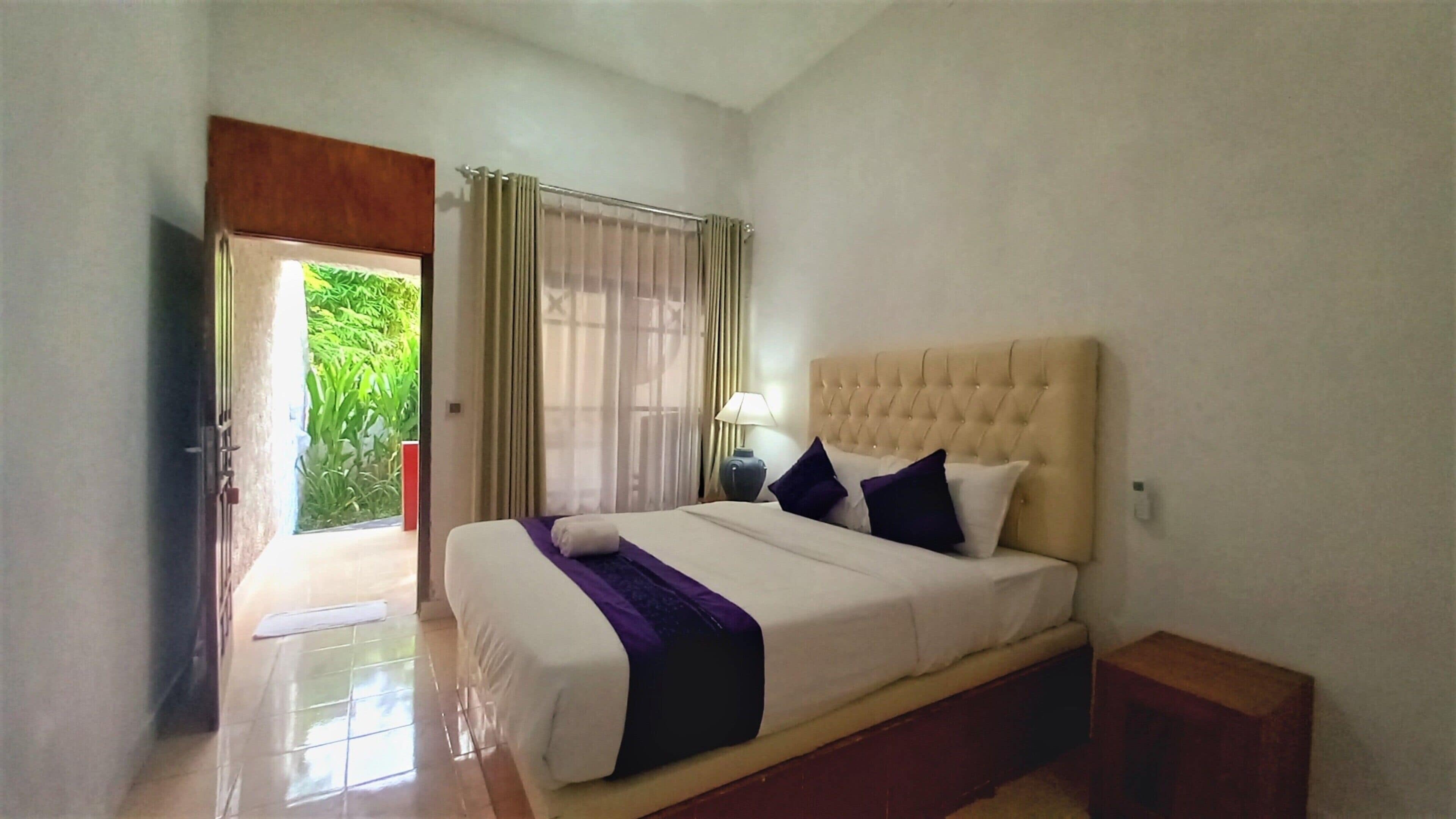 A bedroom with a large bed, a nightstand, a lamp, and a view of greenery through the door.