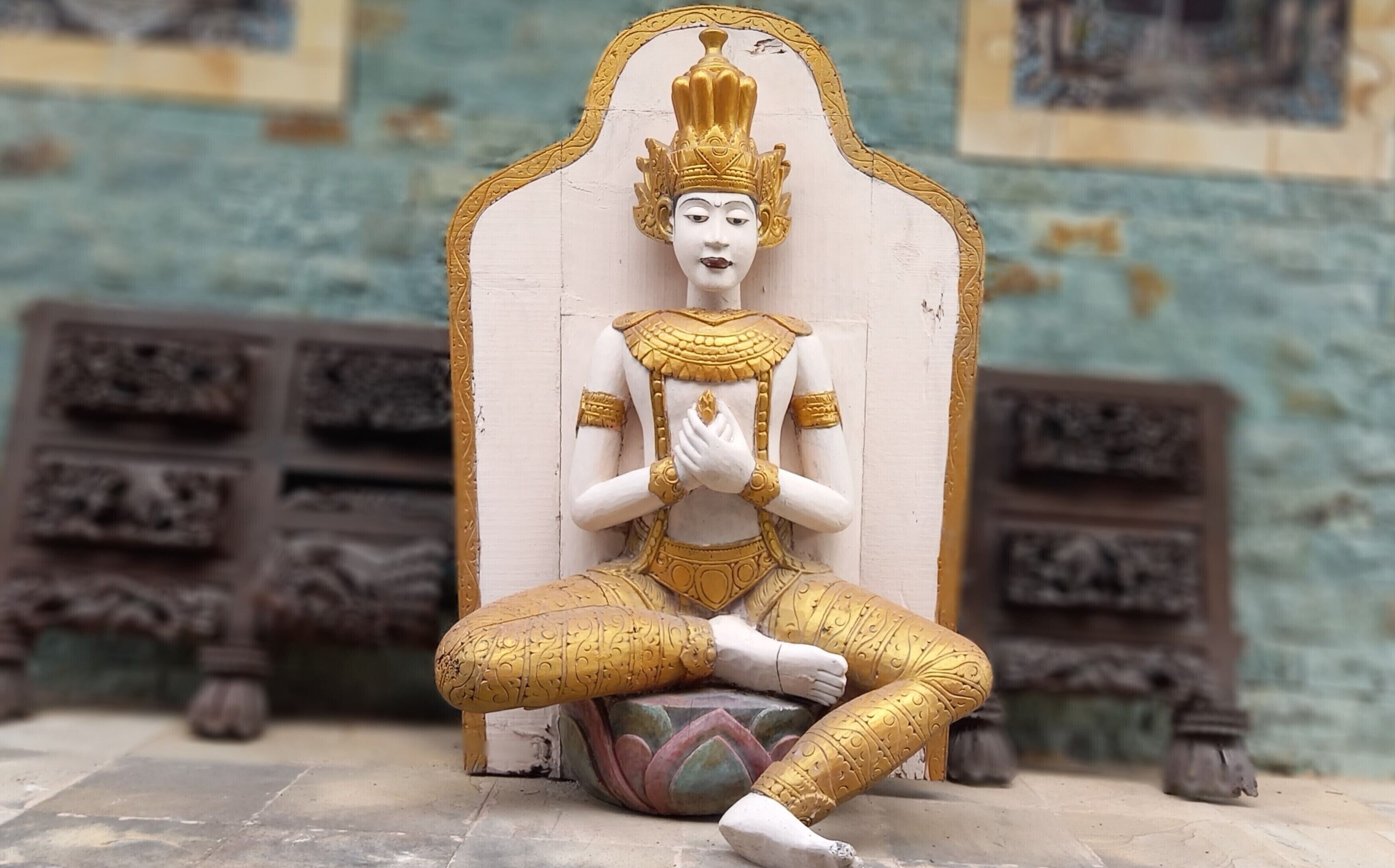 Una statua raffigura una figura seduta in posizione meditativa, con decorazioni in oro elaborate, sullo sfondo di un muro con intagli decorativi.