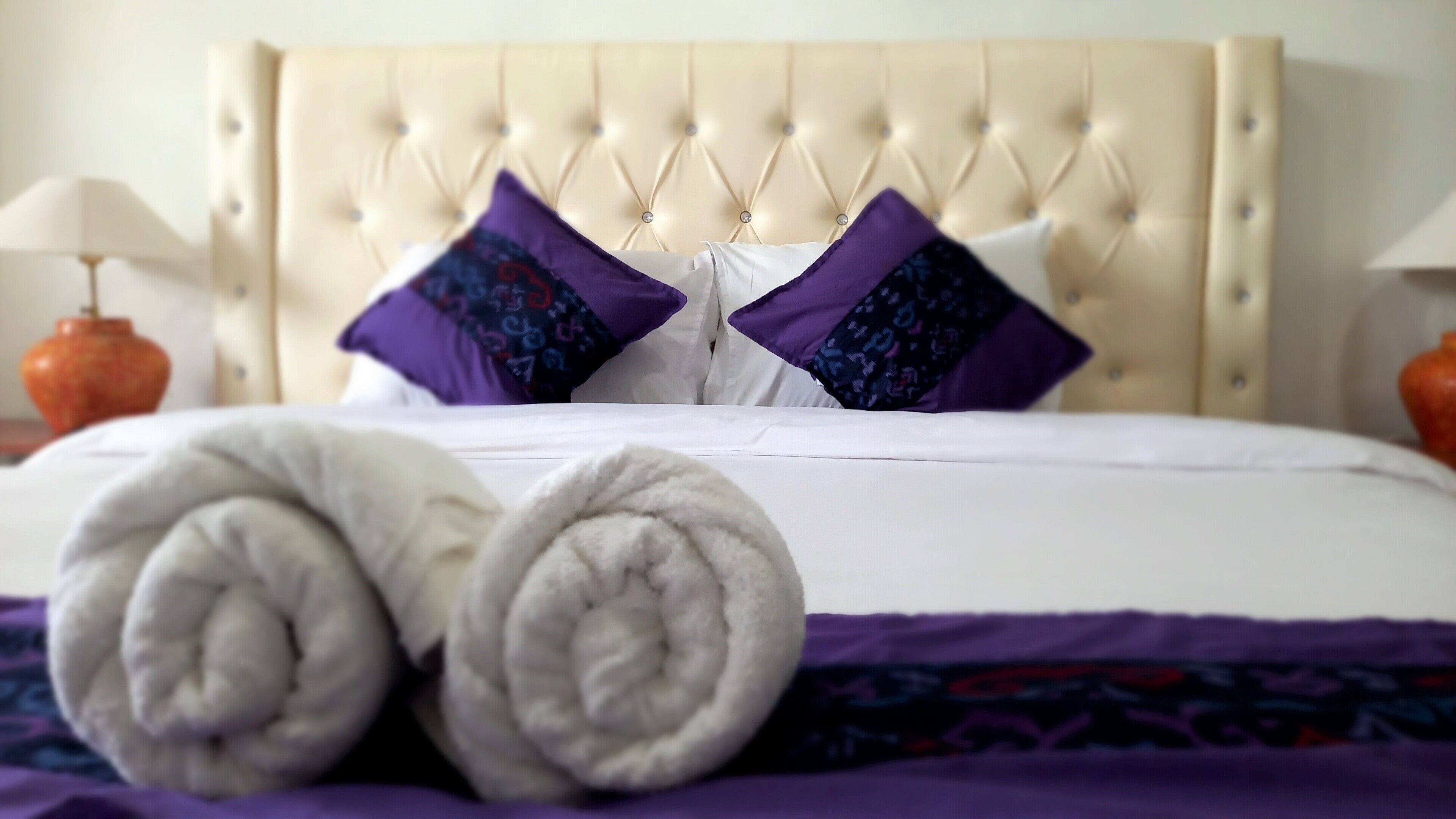 Ein ordentlich gemachtes Bett mit einer gesteppten Kopfstütze, zwei violetten Kissen mit Muster und zwei gerollten weißen Handtüchern auf einer violetten Bettdecke.
