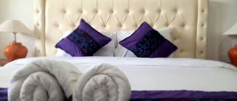 Ein ordentlich gemachtes Bett mit einer gesteppten Kopfstütze, zwei violetten Kissen mit Muster und zwei gerollten weißen Handtüchern auf einer violetten Bettdecke.