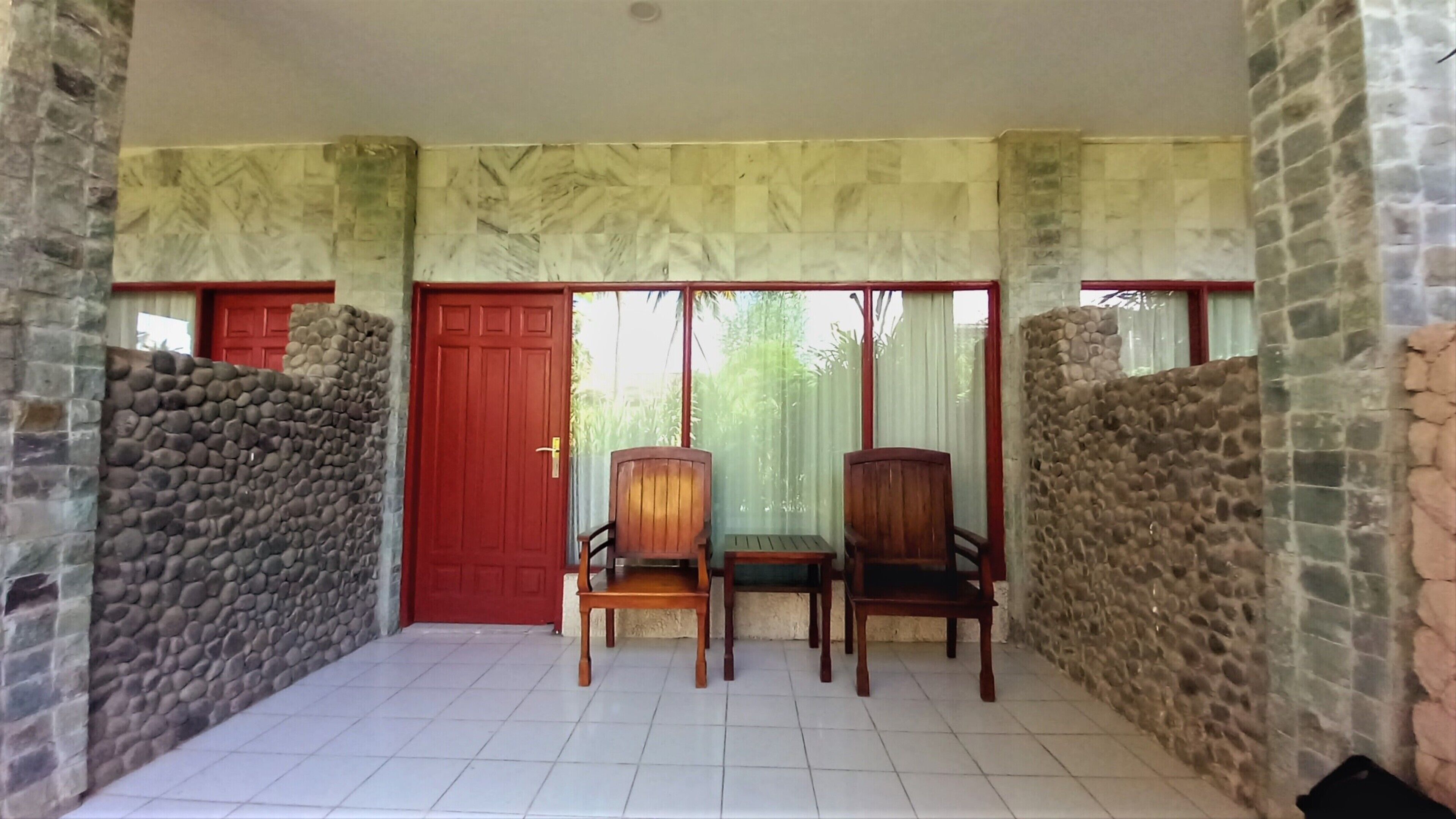 Um quarto com piso de azulejos, porta vermelha, duas cadeiras de madeira e paredes de pedra.