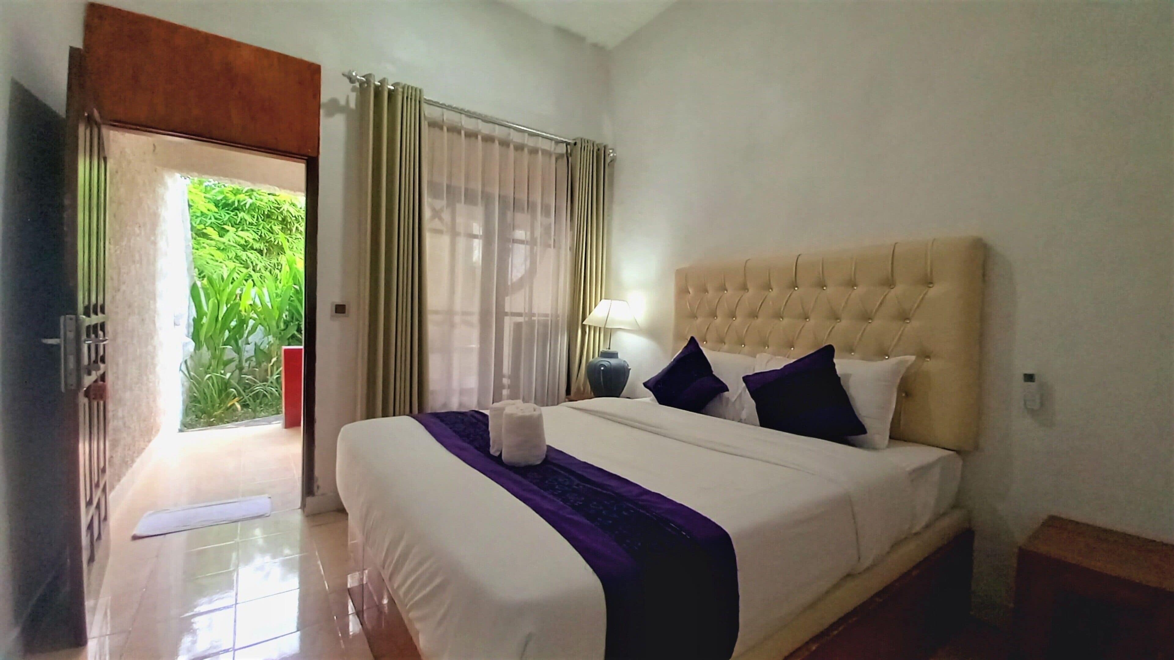 A hotel room with a large bed, a nightstand with a lamp, and a view of greenery through the open door.