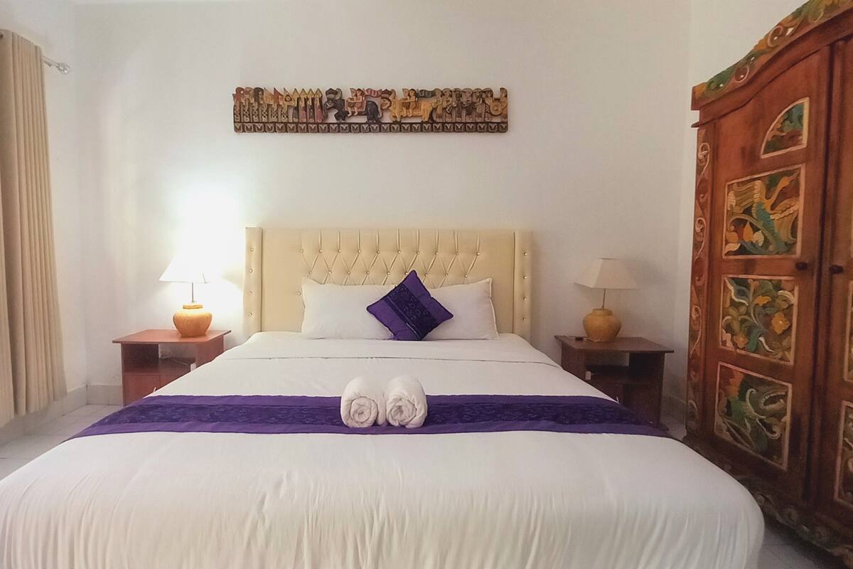 A hotel room with a large bed, two bedside lamps, a decorative headboard, a wall art piece, and a wooden wardrobe.