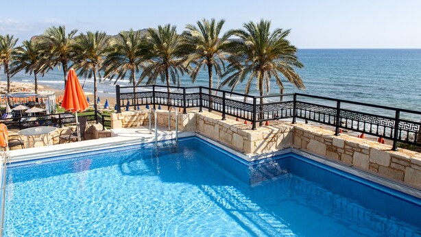 A swimming pool with a black decorative fence, palm trees, and a view of the sea. - Alexandra Beach Hotel (Chania)
