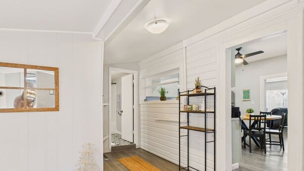 A hallway with white walls, a wooden floor, and a mirror on the left. There is a door leading to a bathroom with a shower and a toilet. A shelf with items is on the right wall. - Single Family Home with Fenced Backyard and AC! (Philomath)