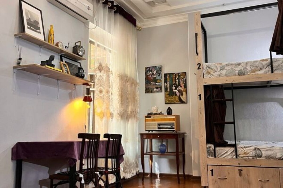 A room with a bunk bed, a dining table with chairs, a small table with a lamp, and shelves with various items.