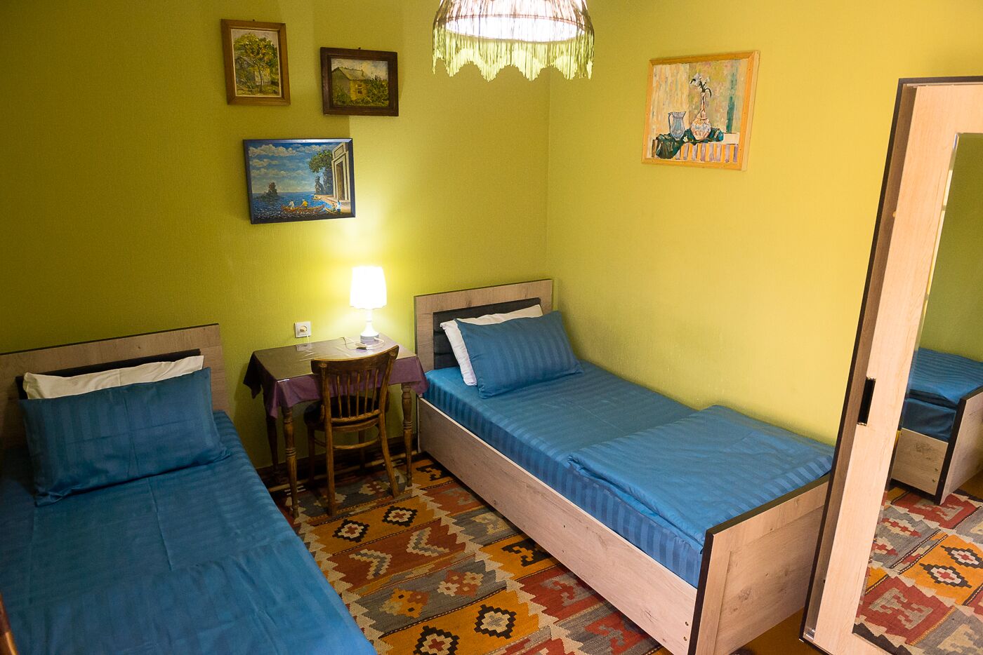 Een kamer met twee eenpersoonsbedden, een kleine tafel met een lamp, een stoel en een kamerkleed met een patroon.