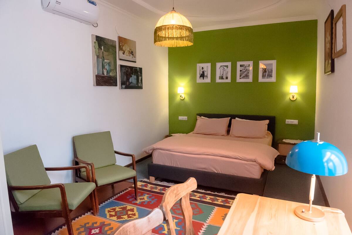 Un dormitorio con una pared verde, una cama con dos almohadas, una silla de madera, un escritorio con una lámpara y luces instaladas en la pared.