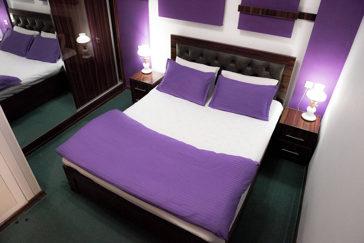 Una cama bien hecha con ropa de cama morada, un cabecero de madera y dos mesitas de noche con lámparas.