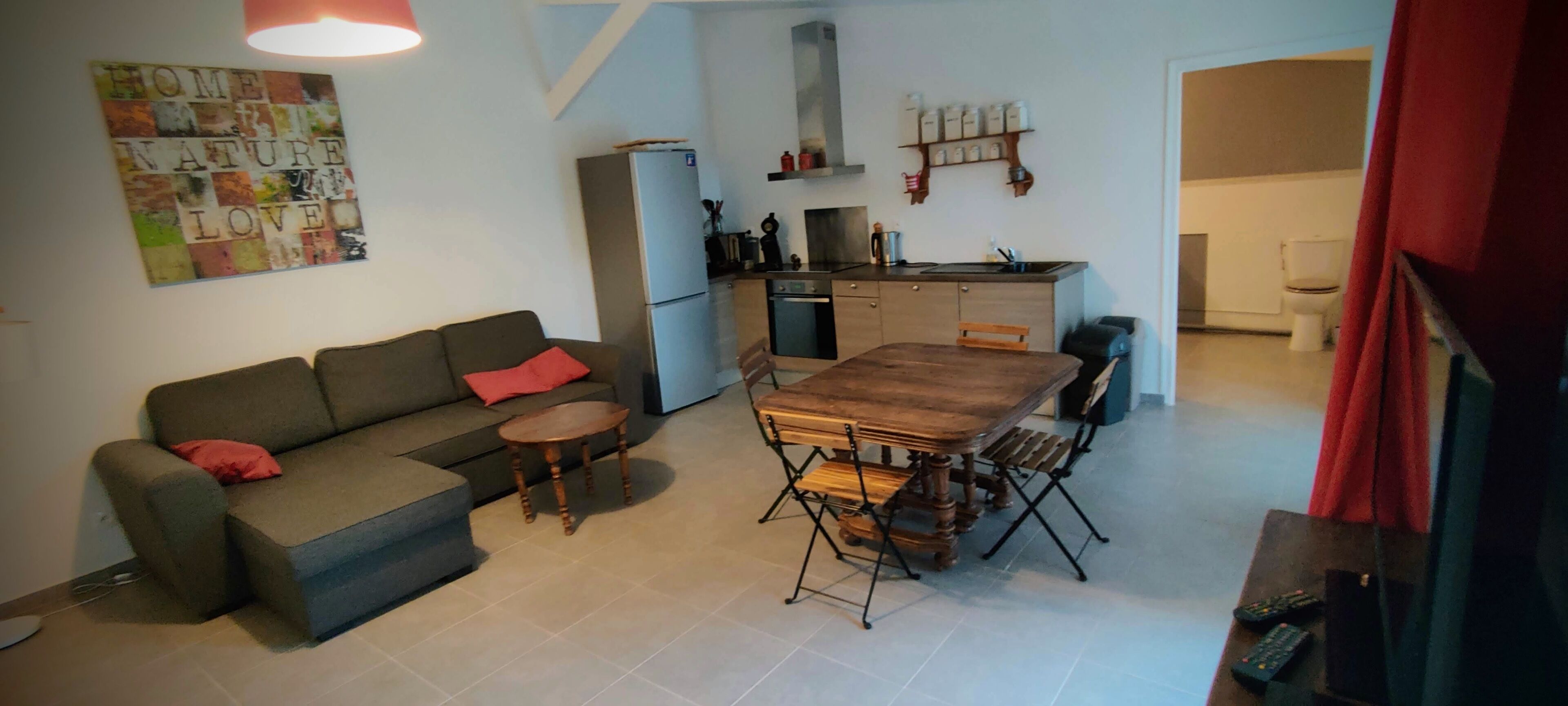 Un ambiente compatto con divano, un piccolo tavolo rotondo, un tavolo da pranzo in legno con sedie, angolo cottura con frigorifero, fornelli ed scaffali, e una tenda rossa parzialmente visibile.