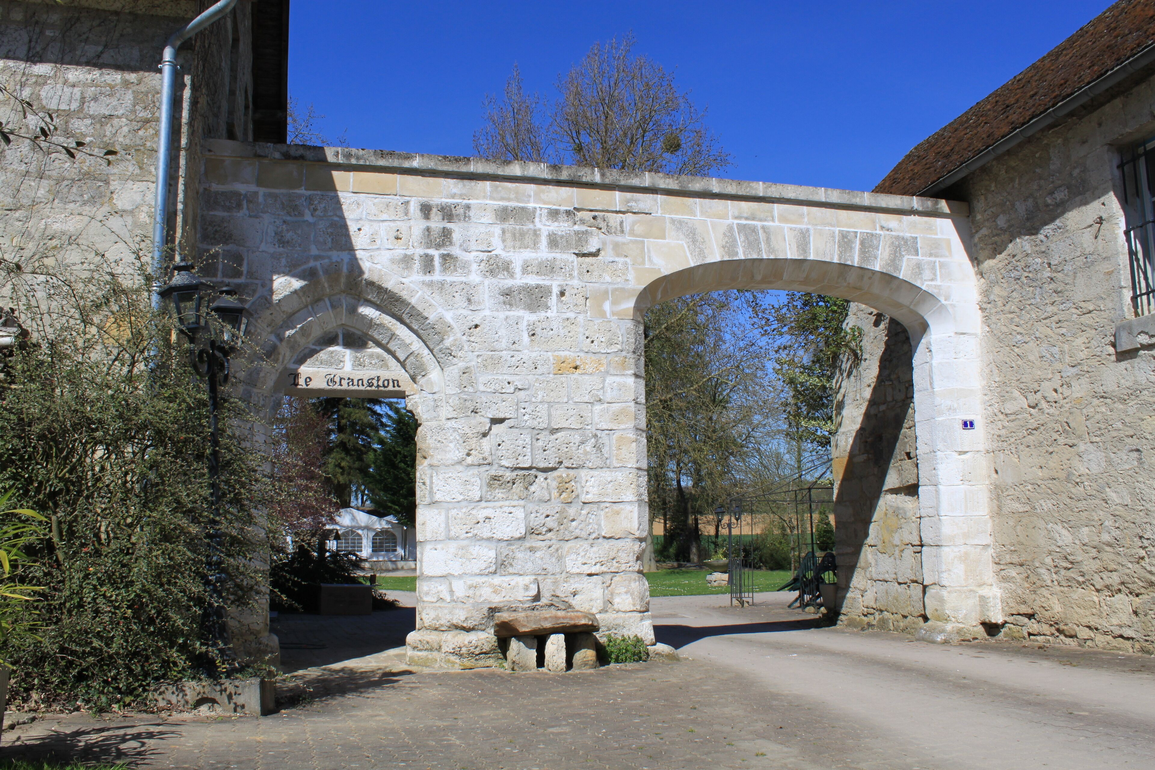 A stone archway with a bench underneath, leading to a building with a sign that reads "Le Grangier."