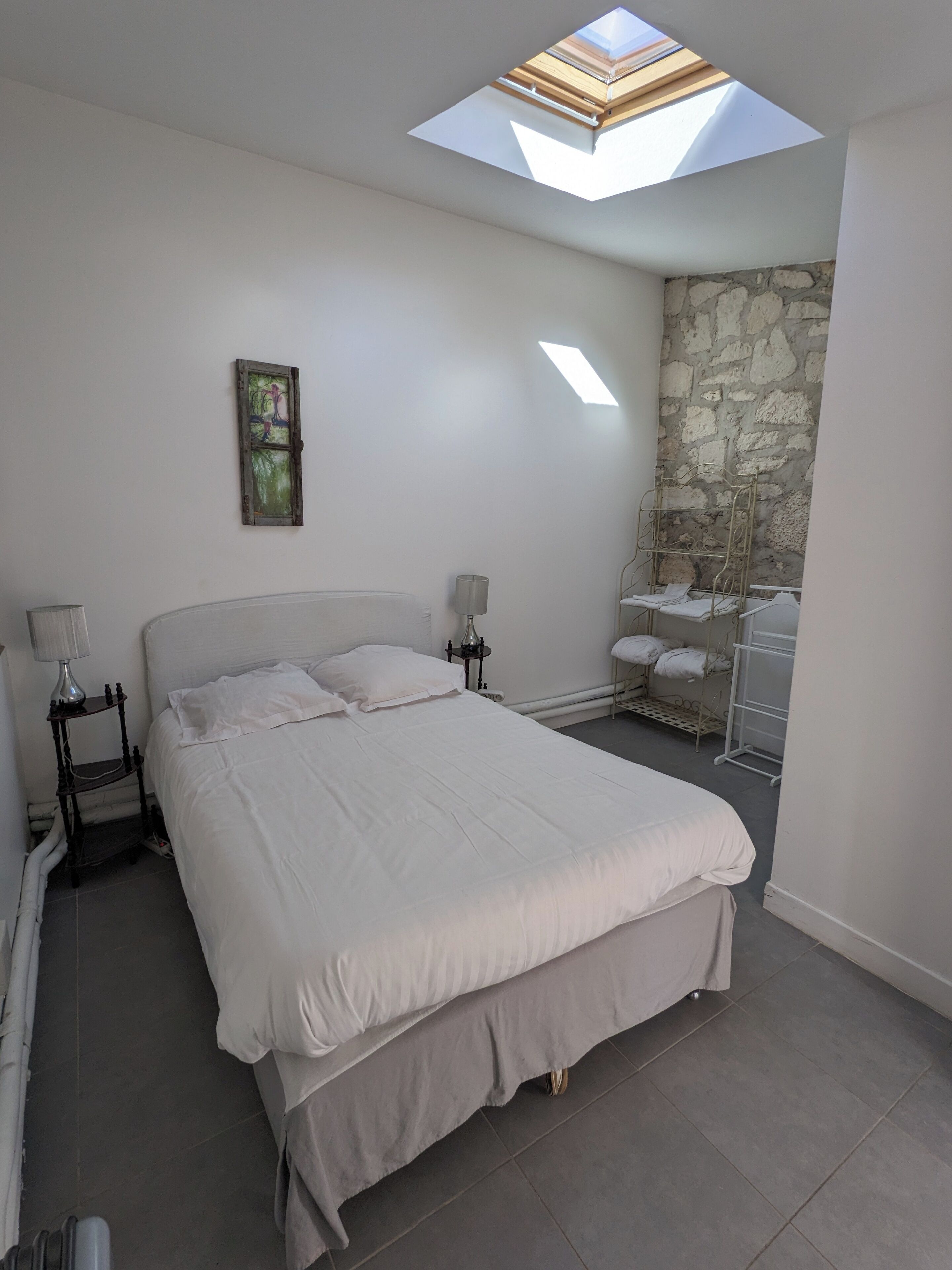Un dormitorio con tragaluz, una cama con ropa blanca, una mesita de noche con lámpara y una pared de piedra.