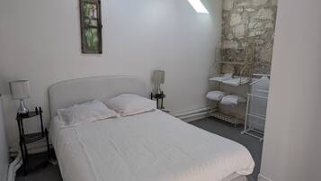 Un dormitorio con tragaluz, una cama con ropa blanca, una mesita de noche con lámpara y una pared de piedra.