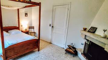 A four-poster bed with a wooden frame, a bedside table with a lamp, a chair, and a fireplace with a mantel.