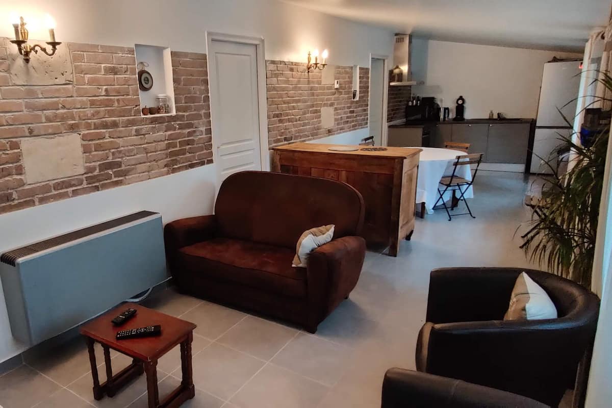 A living room with a brick wall, a brown sofa, a small table with a remote control, and a kitchen in the background.