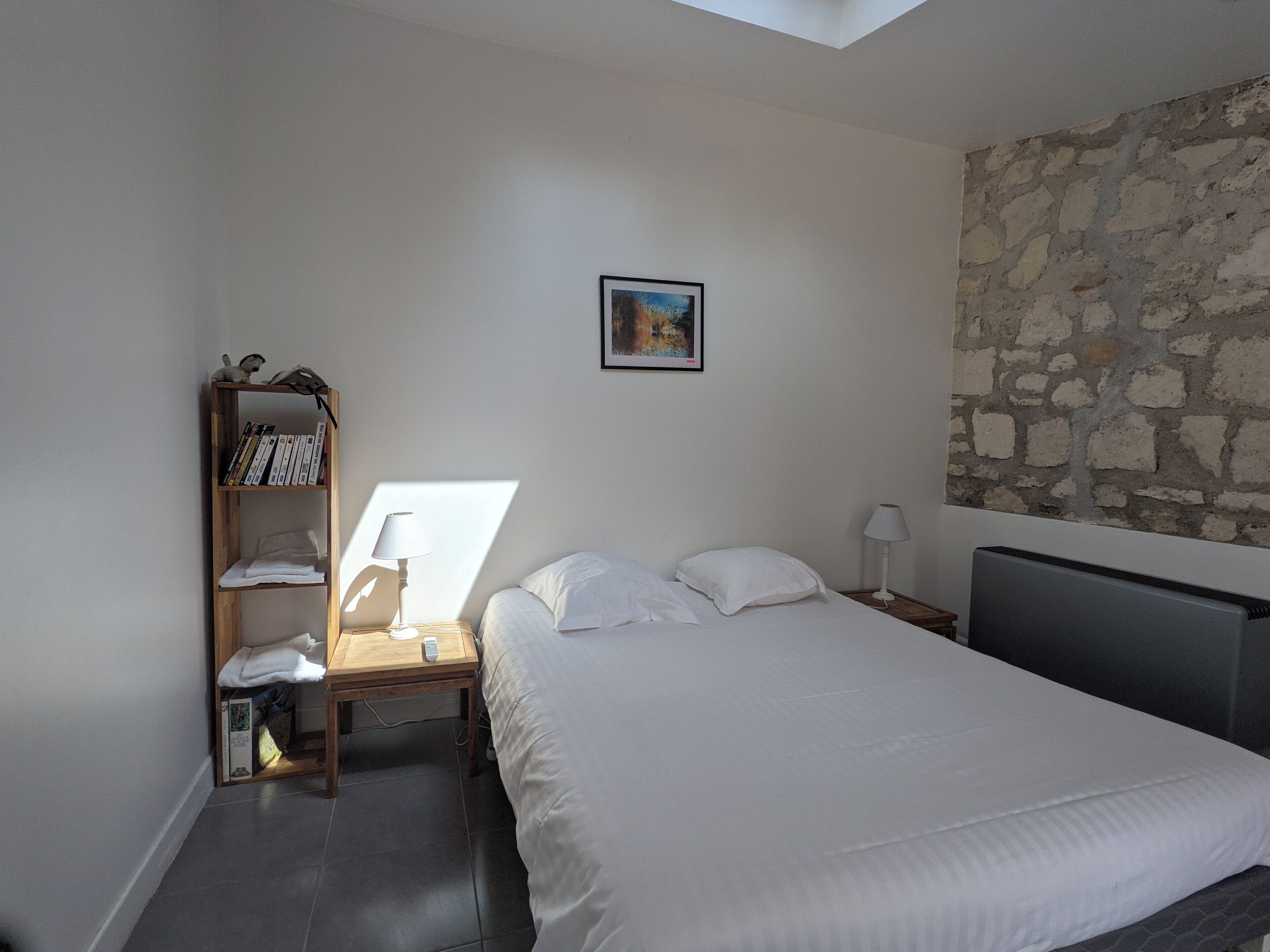 A bedroom with a bed, a nightstand, a bookshelf, a lamp, and a stone wall.