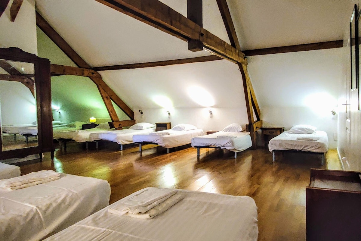 A dormitory-style room with multiple single beds arranged in rows, wooden beams on the ceiling, and a large mirror on one wall.