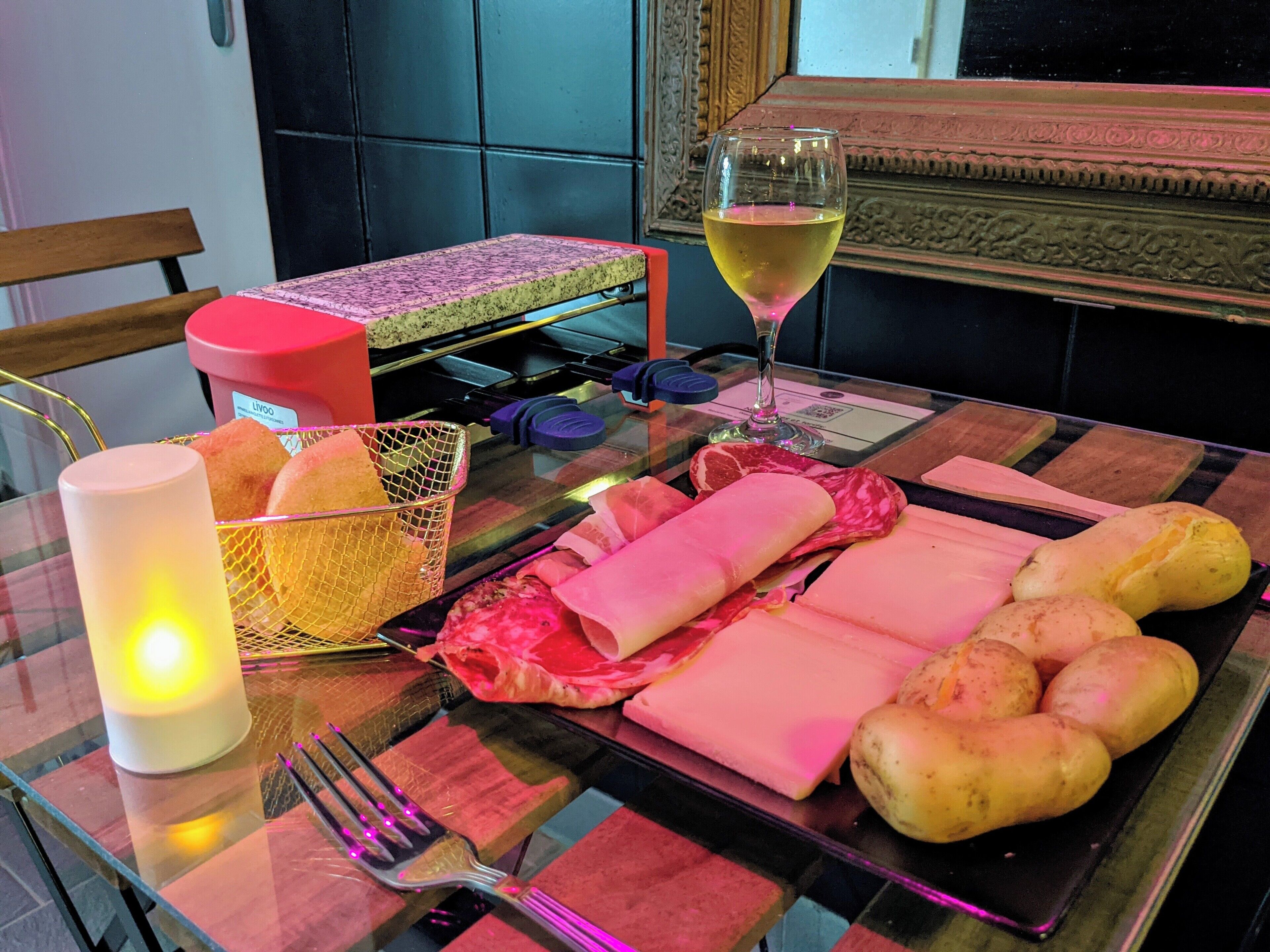 A dining table with a glass top, a candle, a basket of bread, a glass of wine, and various meats and cheeses.