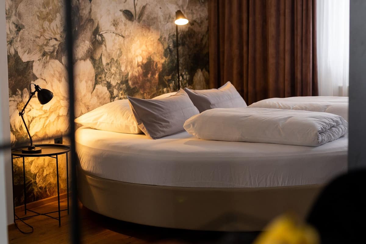 A hotel room with a large bed, a nightstand with a lamp, a floral wall mural, and a window with curtains.