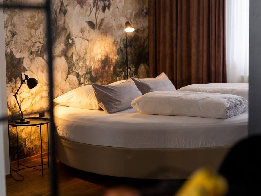 A hotel room with a large bed, a nightstand with a lamp, a floral wall mural, and a window with curtains.