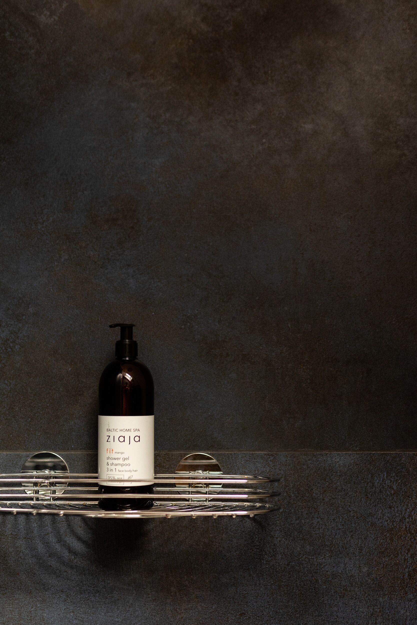 Une salle de bain avec un mur texturé de couleur sombre, un flacon noir de gel douche et de shampoing posé sur un plateau en métal, et un miroir.