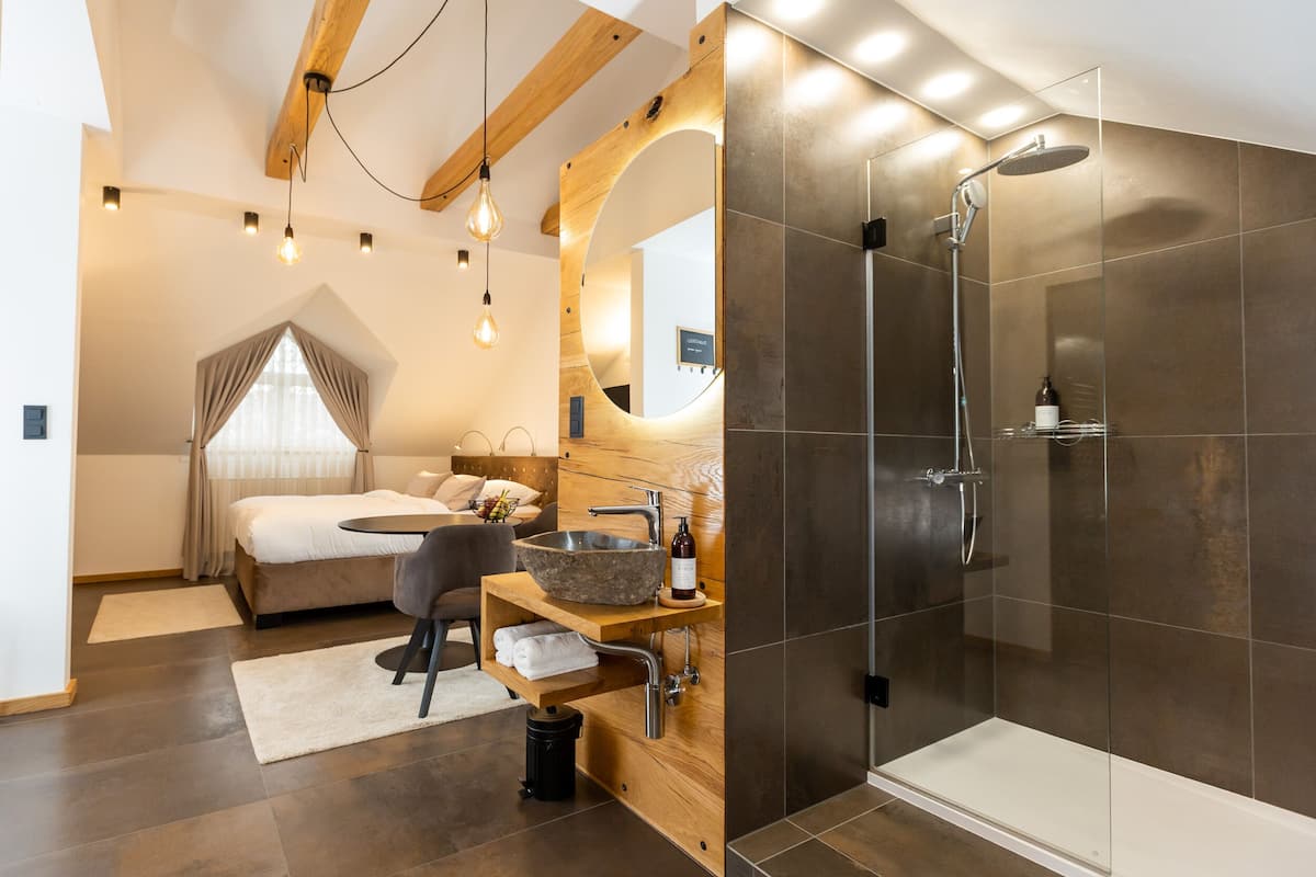 A modern bedroom with a large bed, a round wooden bedside table, a built-in sink, and a shower area with glass walls.