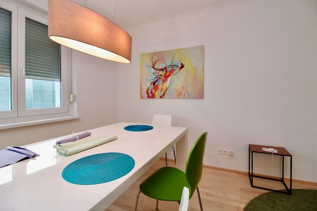 A modern dining area with a white table, green chairs, and a painting of a deer.