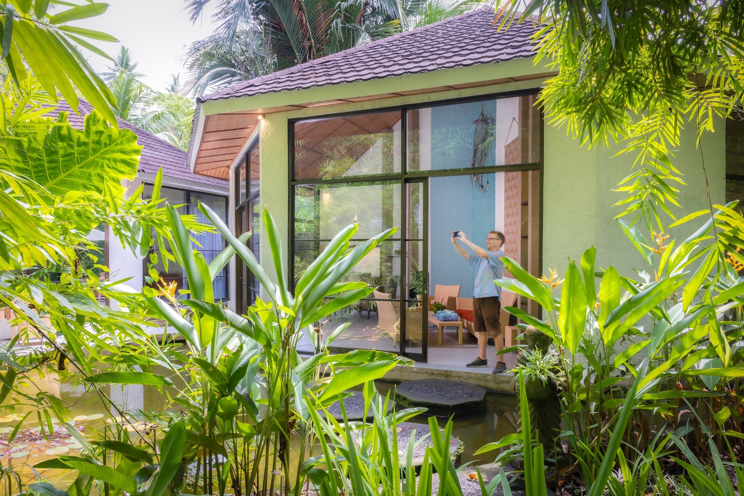 A modern house with large glass windows, surrounded by lush greenery and a small pond.