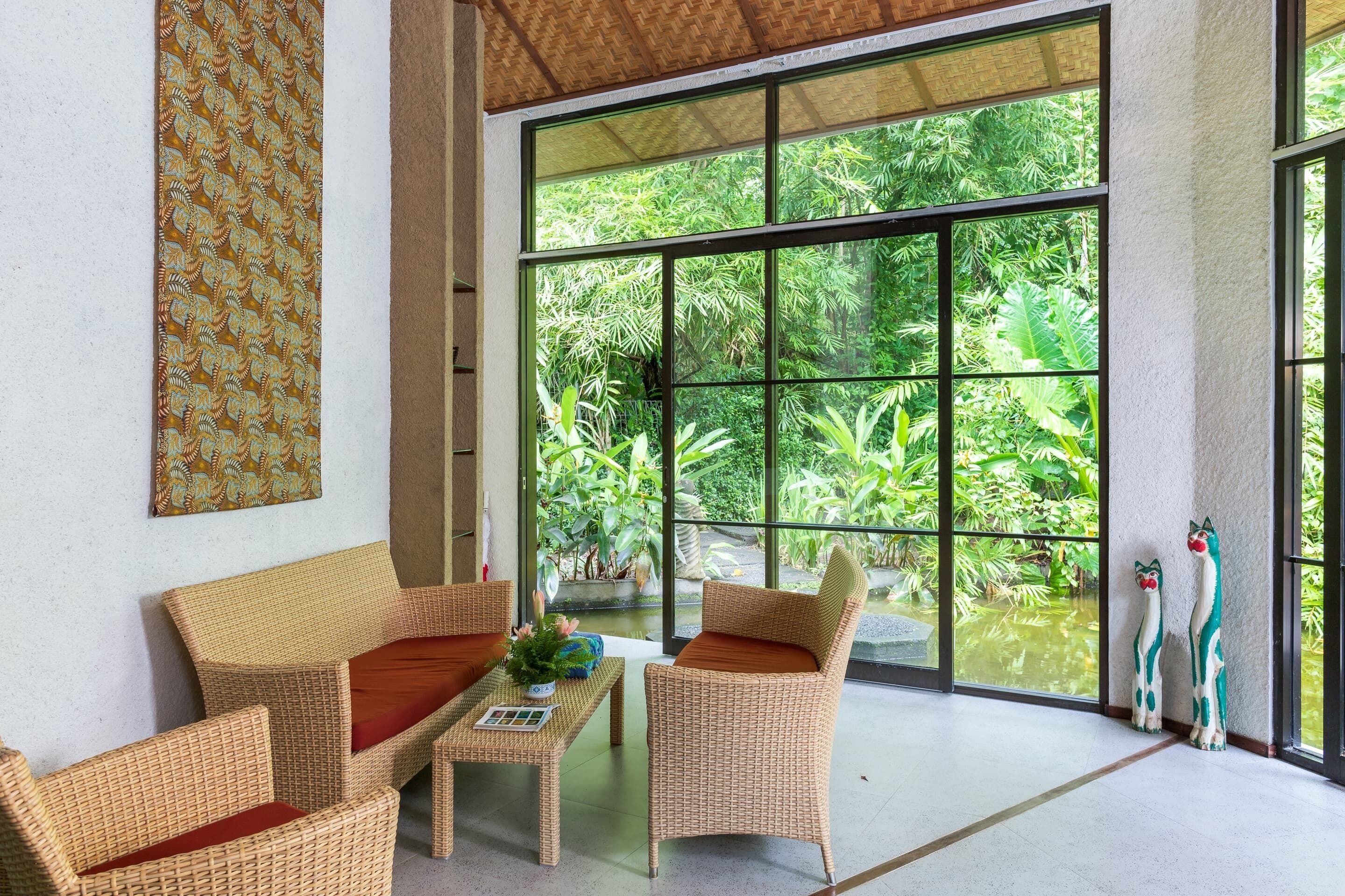 A room with wicker furniture, a large window, and a view of lush greenery.