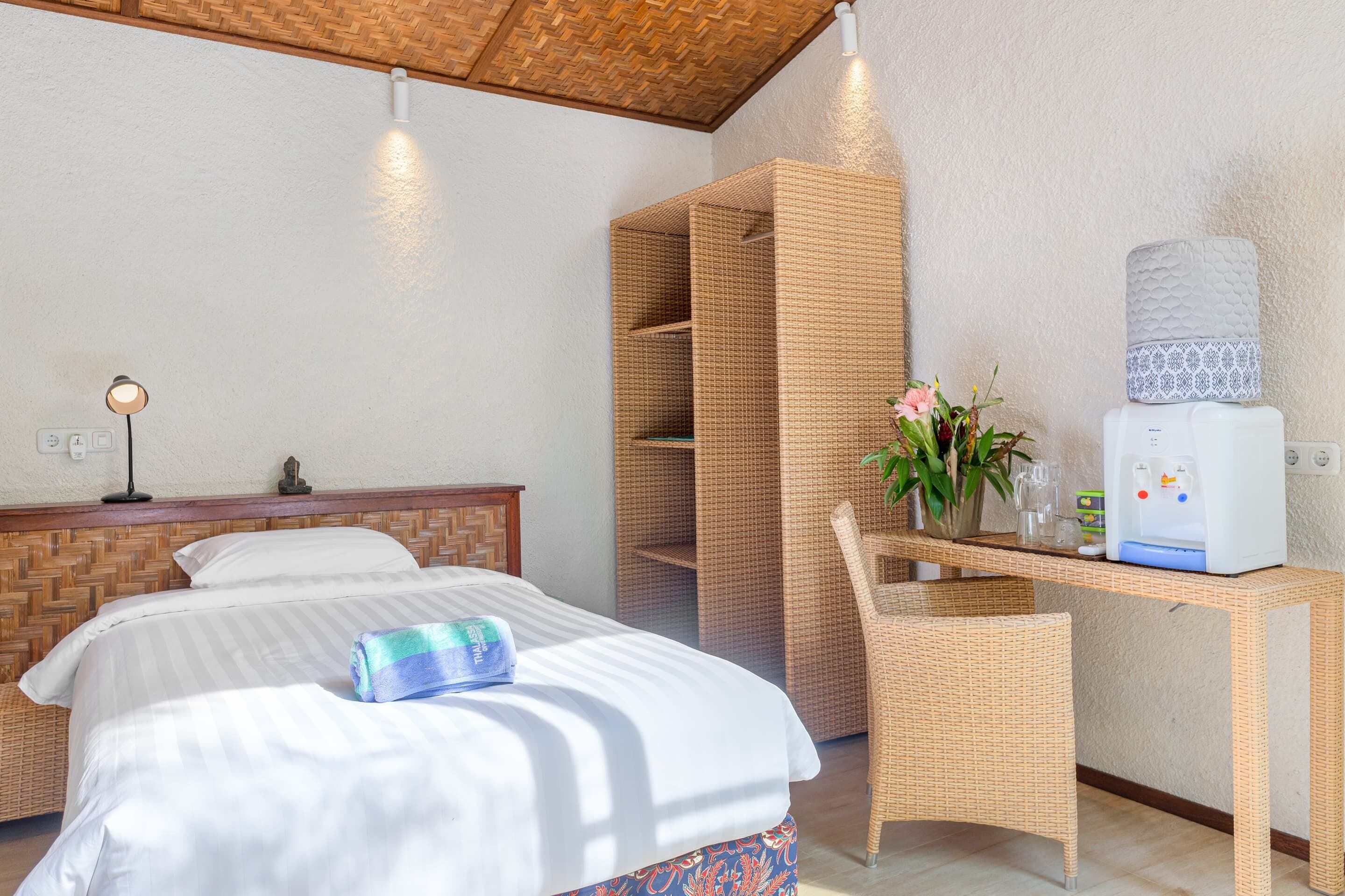 Una camera d'albergo con un letto, una scrivania con dispenser d'acqua, una sedia e una scaffalatura in vimini.