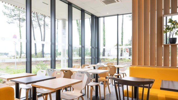 A modern dining area with large windows, wooden tables, and yellow seating. - B&B HOTEL Saint-Geours-De-Maremne Hossegor (Saint-Geours-de-Maremne)