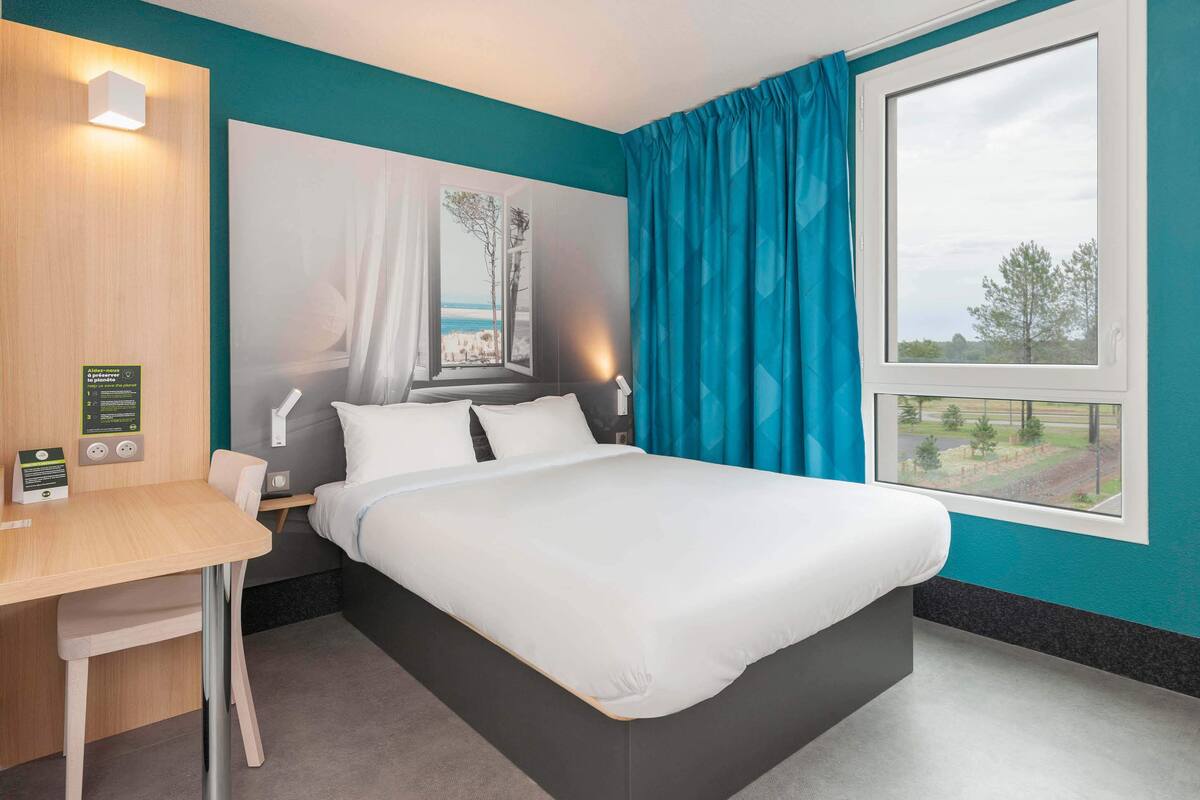 Quarto de hotel com cama, escrivaninha, janela com cortinas e vista para o exterior.