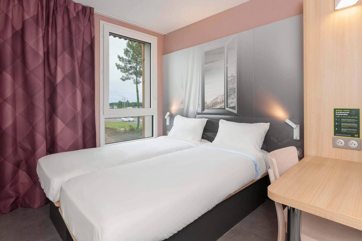 Quarto de hotel com uma cama, janela, luminária fixada na parede e uma pequena mesa.