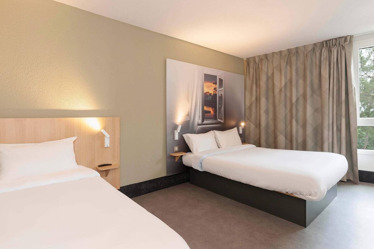 Quarto de hotel com duas camas, uma janela grande com cortinas, uma luminária na parede e um quadro.