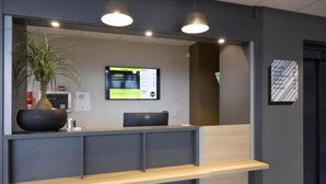 A reception desk with a computer, a potted plant, and a digital information board. - B&B HOTEL Saint-Geours-De-Maremne Hossegor (Saint-Geours-de-Maremne)