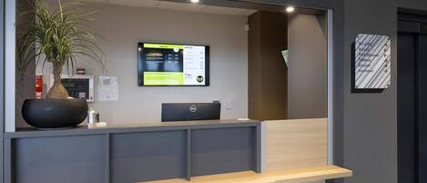 A reception desk with a computer, a potted plant, and a digital information board.