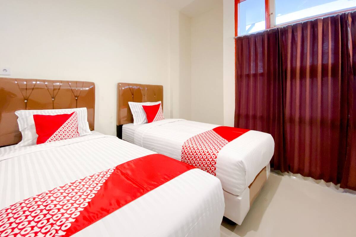 A hotel room with two beds, each with white bedding and red accents, a wooden headboard, and a window with red curtains.