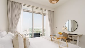 A hotel room with a large bed, a desk with a chair, a round mirror, and a view of the city through large windows. - Maison Privee - Graceful Apt in Prestigious Dubai Hills close to the Golf Course (Dubai)