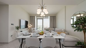 A modern dining area with a white table set for six, surrounded by grey chairs. A chandelier hangs above, and a large mirror reflects part of the room. - Maison Privee - Graceful Apt in Prestigious Dubai Hills close to the Golf Course (Dubai)