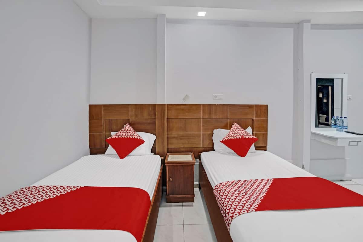 A hotel room with two single beds, each with a wooden headboard, white bedding, and red pillows. There is a small wooden nightstand between the beds, and a white sink with a mirror in the corner.