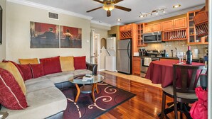 A living room with a sofa, coffee table, and a dining area with a table and chairs. A kitchen with wooden cabinets and appliances is visible in the background. - Romance in the City - 1 Bdrm Studio in Downtown! (St. Petersburg)