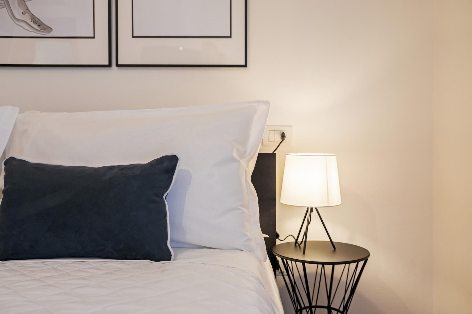 A neatly made bed with white and dark pillows, a round table with a lamp, and framed artwork on the wall.