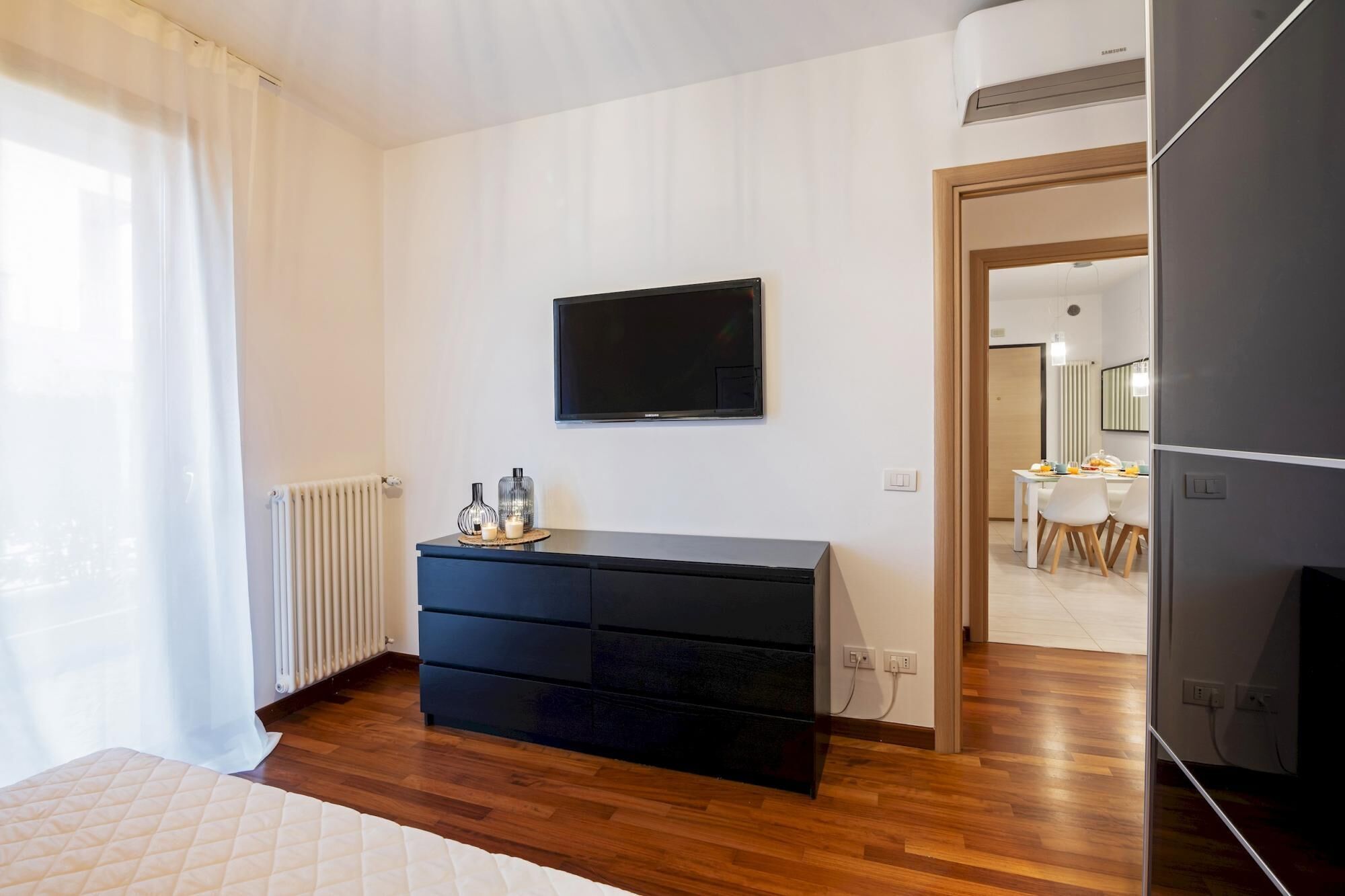 Ein modernes Schlafzimmer mit Holzboden, einer schwarzen Kommode, einem Flachbildfernseher und einer Heizung.