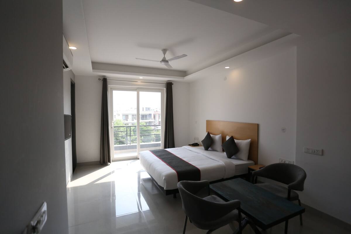 Habitación de hotel moderna con una cama grande, un escritorio y una silla. Cuenta con una ventana con cortinas, un ventilador de techo y vistas a edificios.