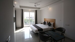 A modern hotel room with a large bed, a desk, and a chair. There is a window with curtains, a ceiling fan, and a view of buildings outside.