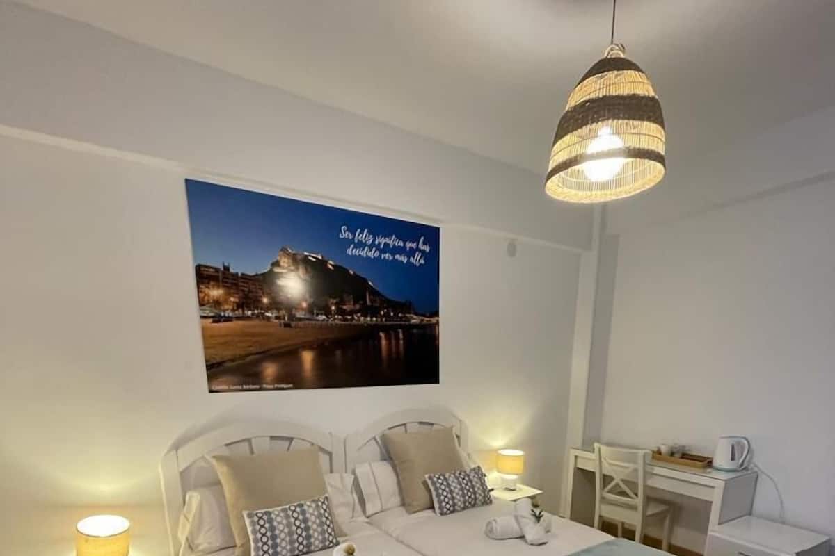 Een hotelkamer met twee bedden, een bureau, een stoel, een nachtkastje met een lamp en een aan de muur hangend stadsbeeld bij nacht.