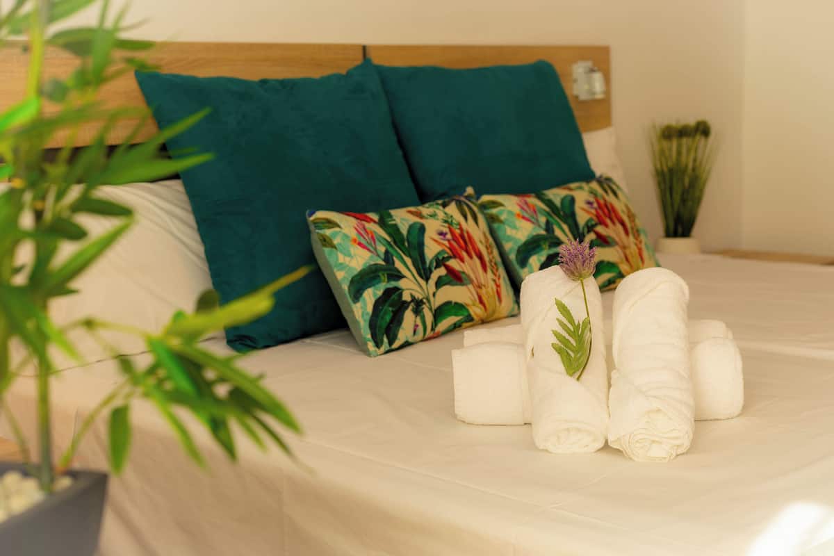 Een net opgemaakt bed met een bloemenkussen, een bloempot en opgerolde handdoeken.