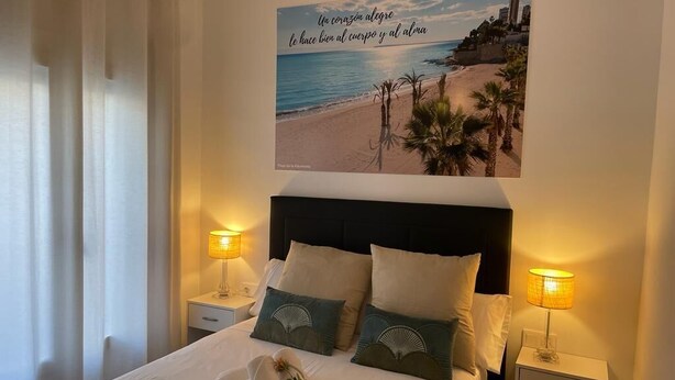 A neatly made bed with pillows and a towel, a bedside table with a lamp, and a wall with a beach scene and text. - Hostal Ventura Premium  (Alicante)